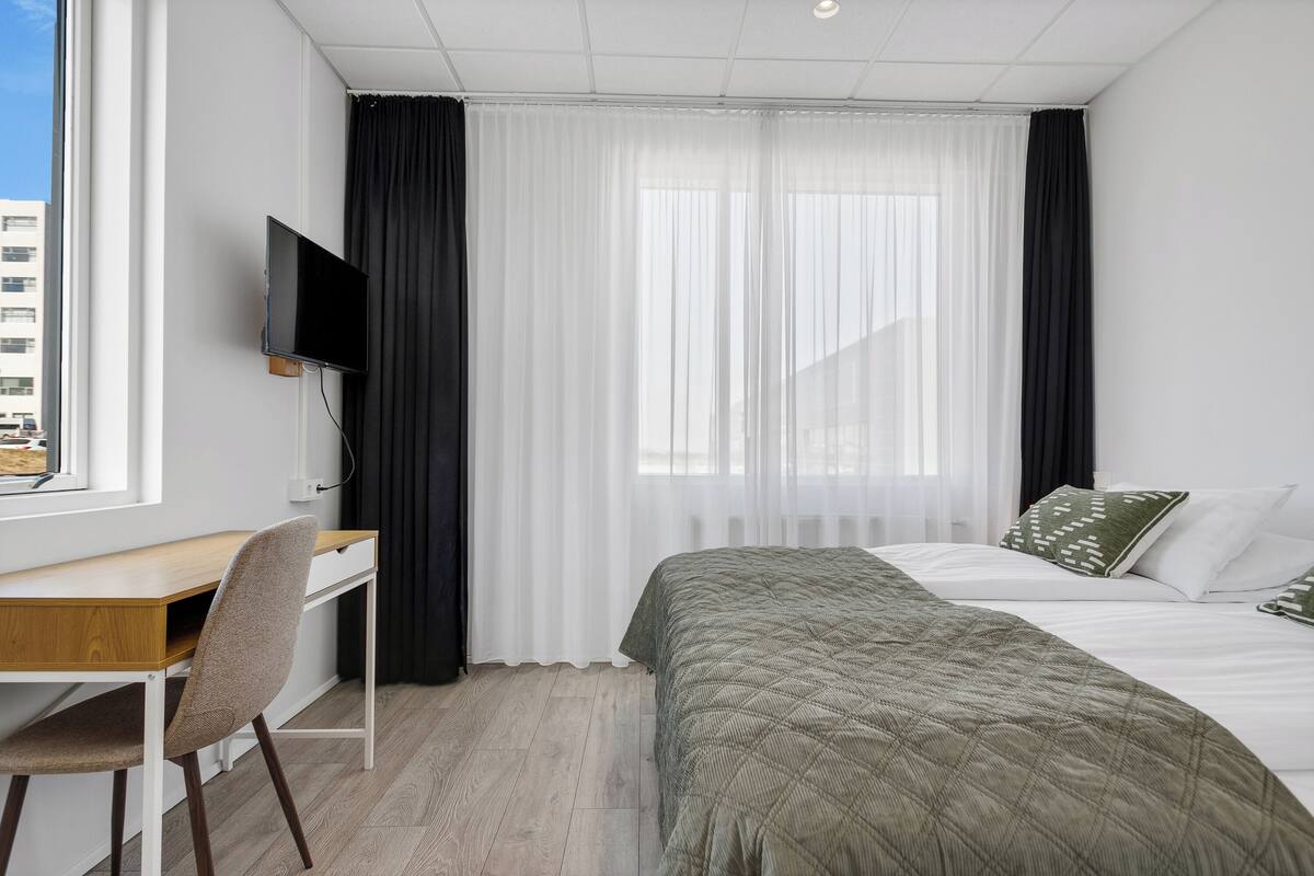 A hotel room with a bed, a desk, a chair, a TV, and sheer curtains.