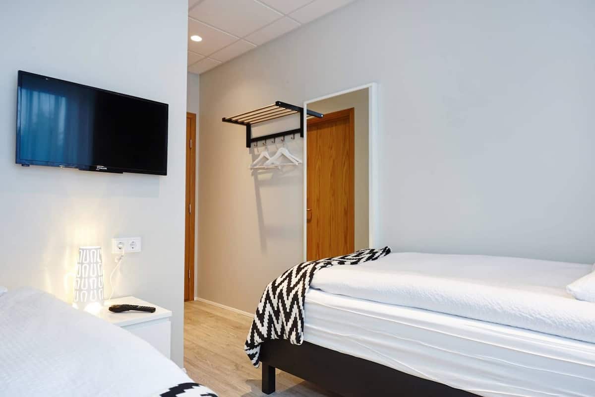 A hotel room with a TV, a bedside table with a lamp, a bench, and a wardrobe with a hanging shelf.