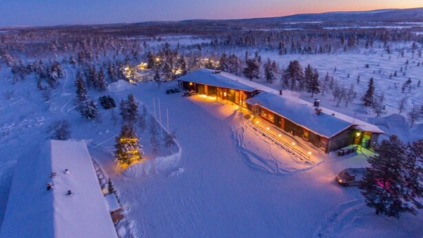 Una cabaña nevada con un edificio central grande y construcciones más pequeñas, rodeada de árboles y un paisaje cubierto de nieve.