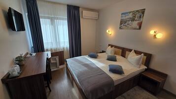A hotel room with a bed, bedside tables, a desk, a chair, a flat-screen TV, a window with curtains, an air conditioning unit, and wall-mounted lights.