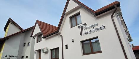 A two-story building with a sign reading "Pensionia Transania." It features a red-tiled roof, multiple windows, and an air conditioning unit on the exterior wall.