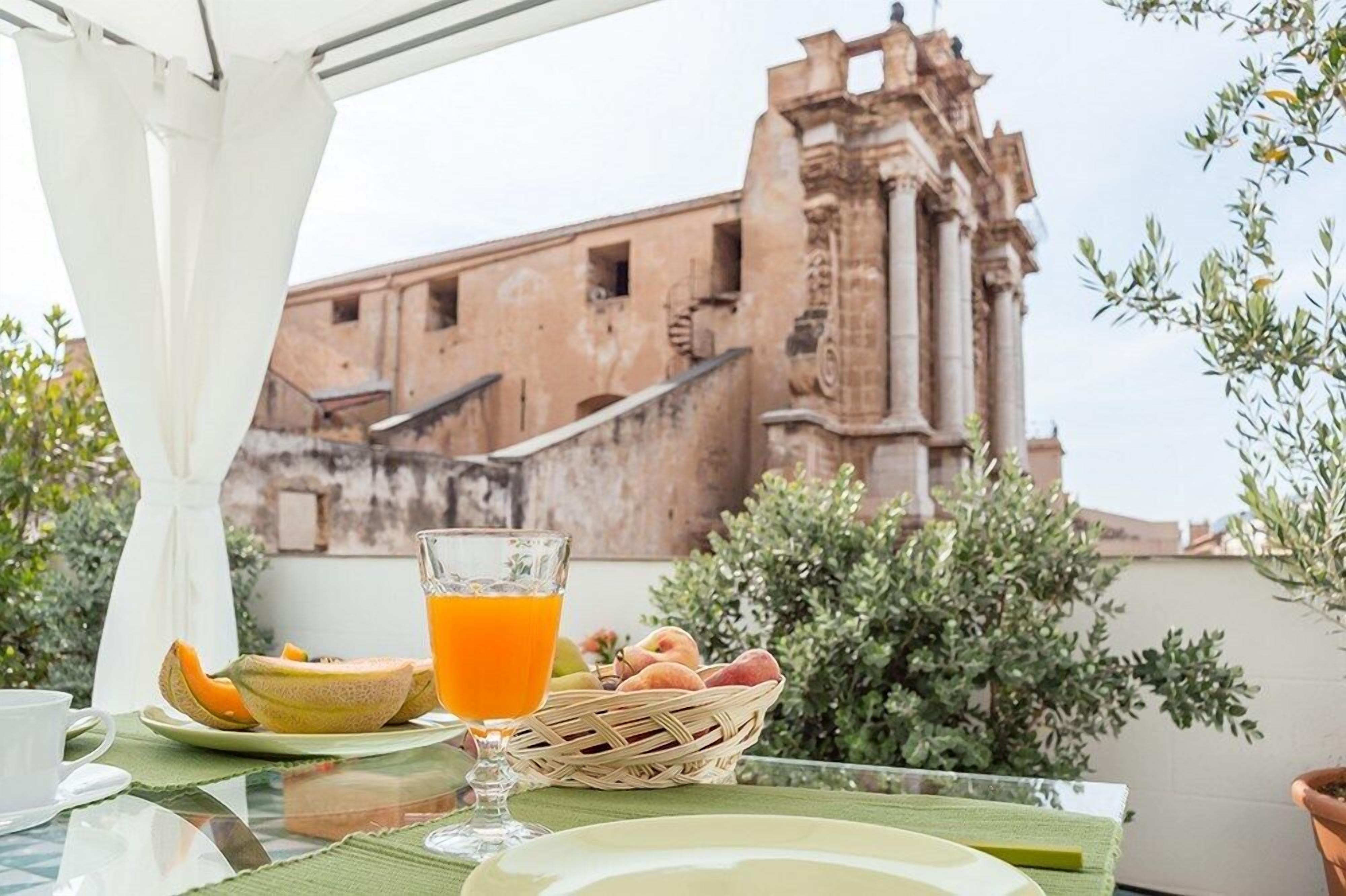 A breakfast setup with a glass of orange juice, melon, and a basket of fruits on a balcony with a view of a historic building.