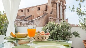 Mesa de café da manhã com copo de suco de laranja, melão e cesta de frutas em uma varanda com vista para um prédio histórico.