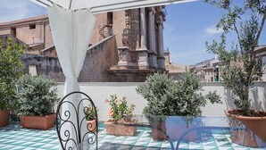 Um terraço com chão xadrez, plantas em vasos, uma cadeira de metal e vista para arquitetura histórica.