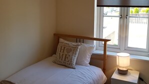 A bedroom with a bed, a bedside table with a lamp, a window with a view, and a pillow with text. - Livingston 4 Bedroom Townhouse (Livingston)