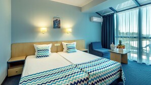 Une chambre d’hôtel avec un grand lit, deux tables de chevet, un bureau avec un panier de fruits, et une vue sur l’extérieur grâce à de grandes fenêtres.