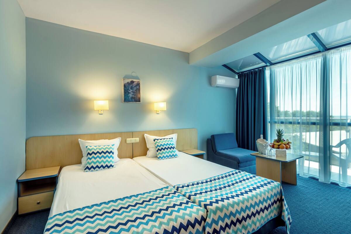 Une chambre d’hôtel avec un grand lit, deux tables de chevet, un bureau avec un panier de fruits, et une vue sur l’extérieur grâce à de grandes fenêtres.