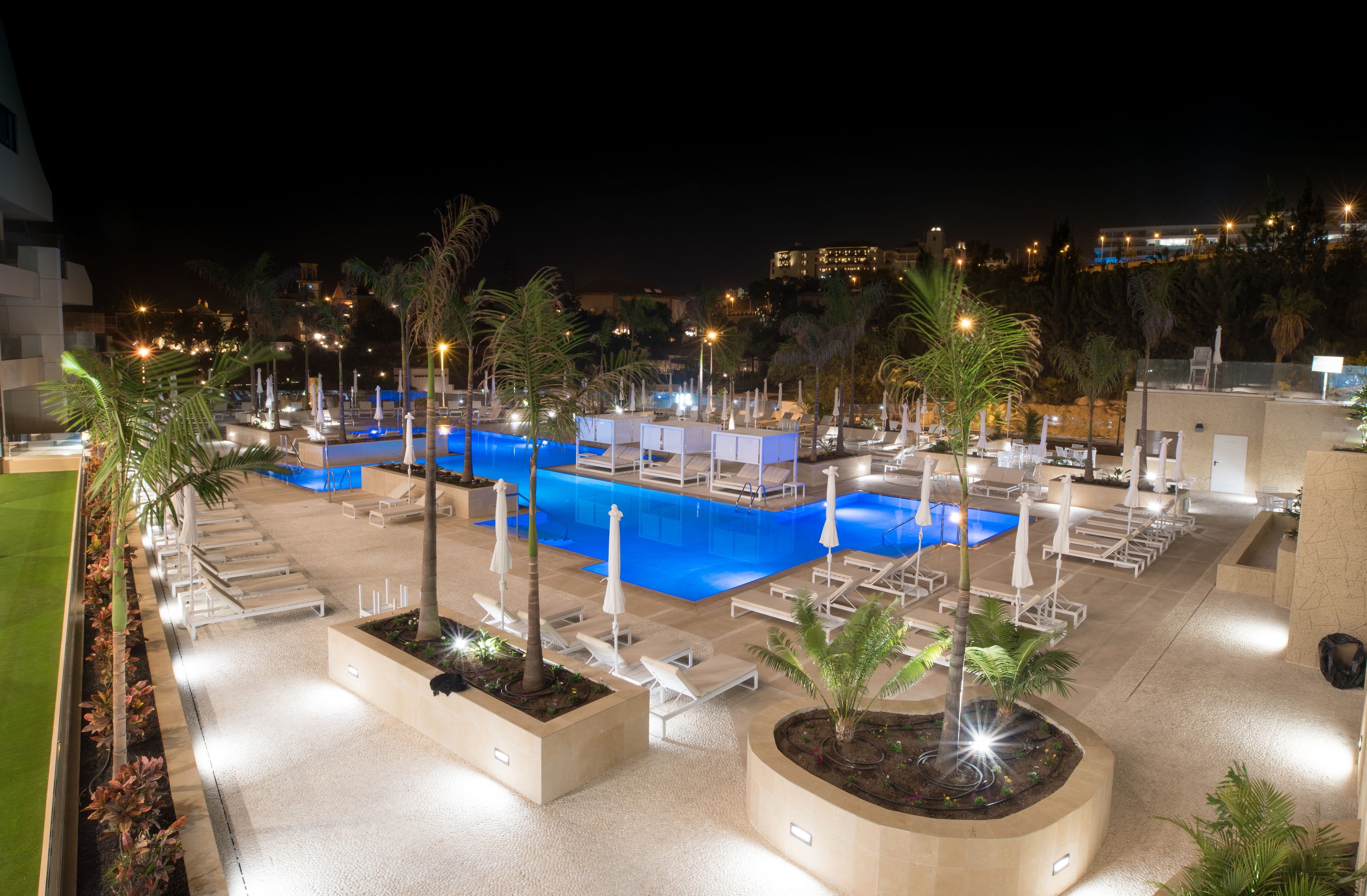A nighttime view of a hotel pool area with multiple lounge chairs and umbrellas, surrounded by palm trees and well-lit by artificial lights.