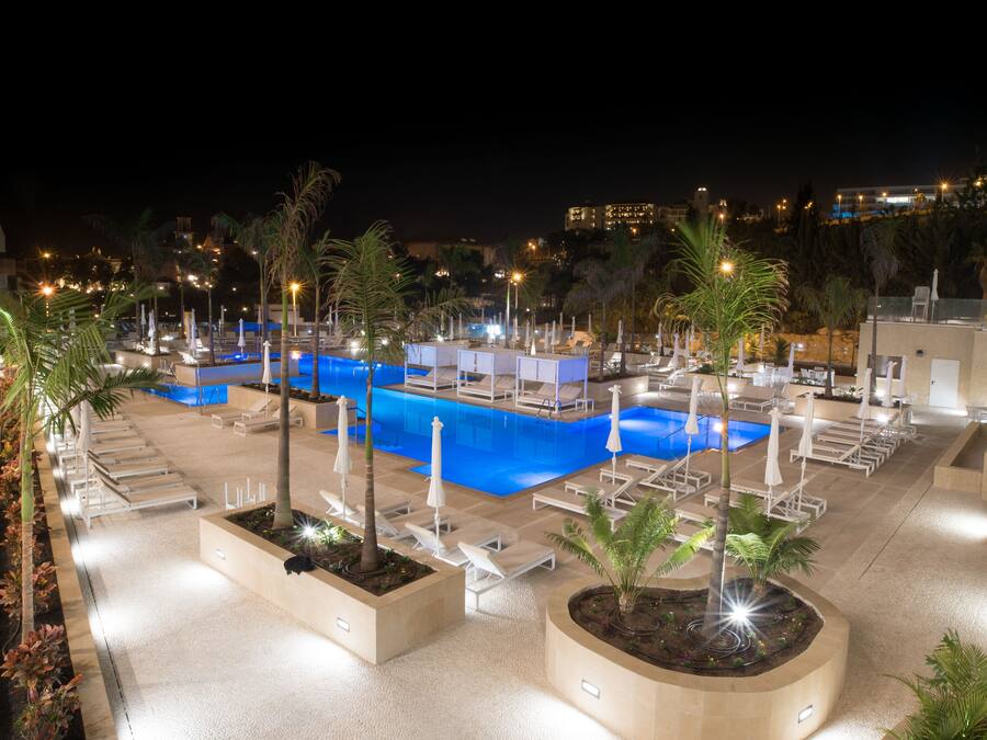 A nighttime view of a hotel pool area with multiple lounge chairs and umbrellas, surrounded by palm trees and well-lit by artificial lights.