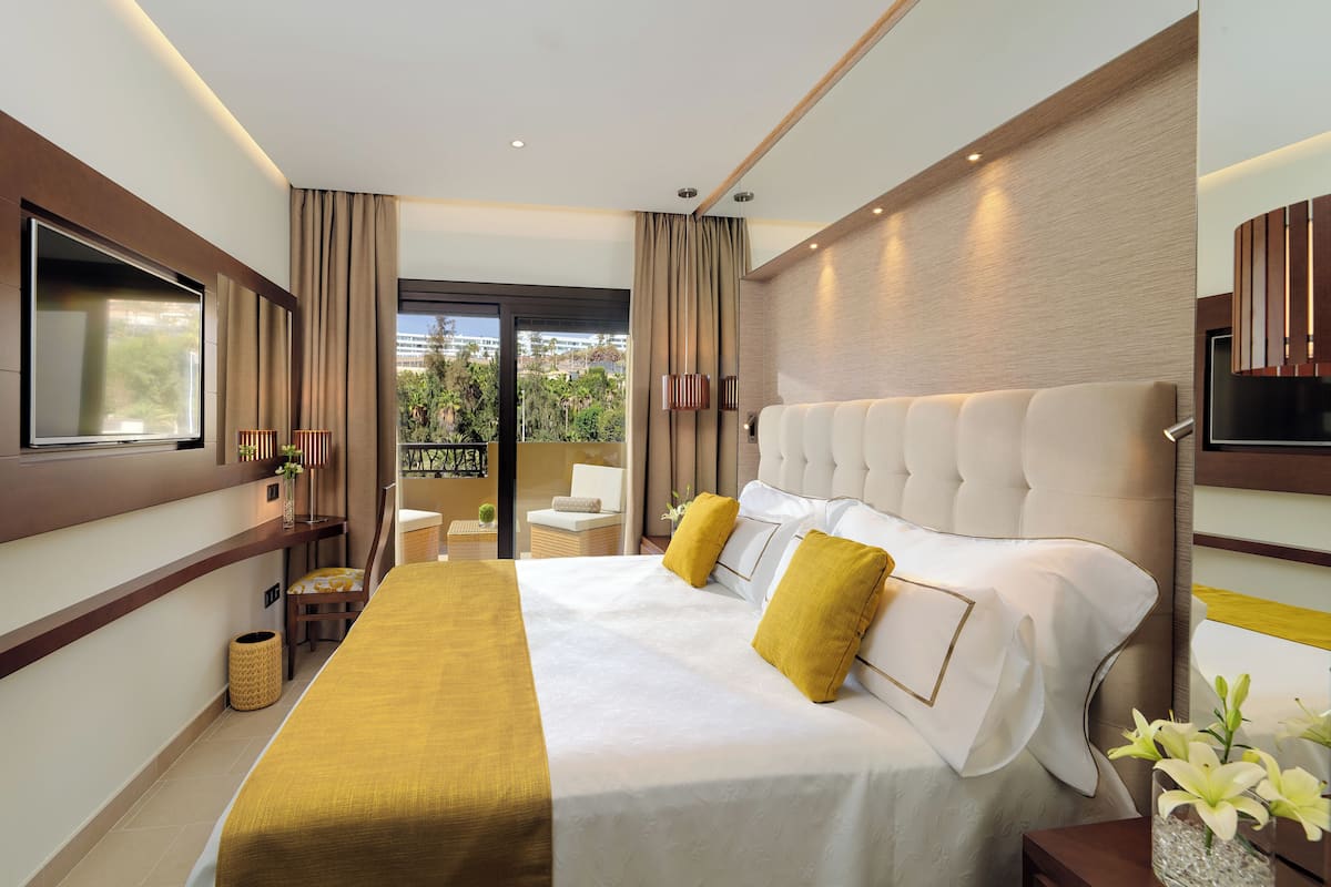 A modern hotel room with a large bed, a television, a balcony with a view of greenery, and a small table with a lamp.