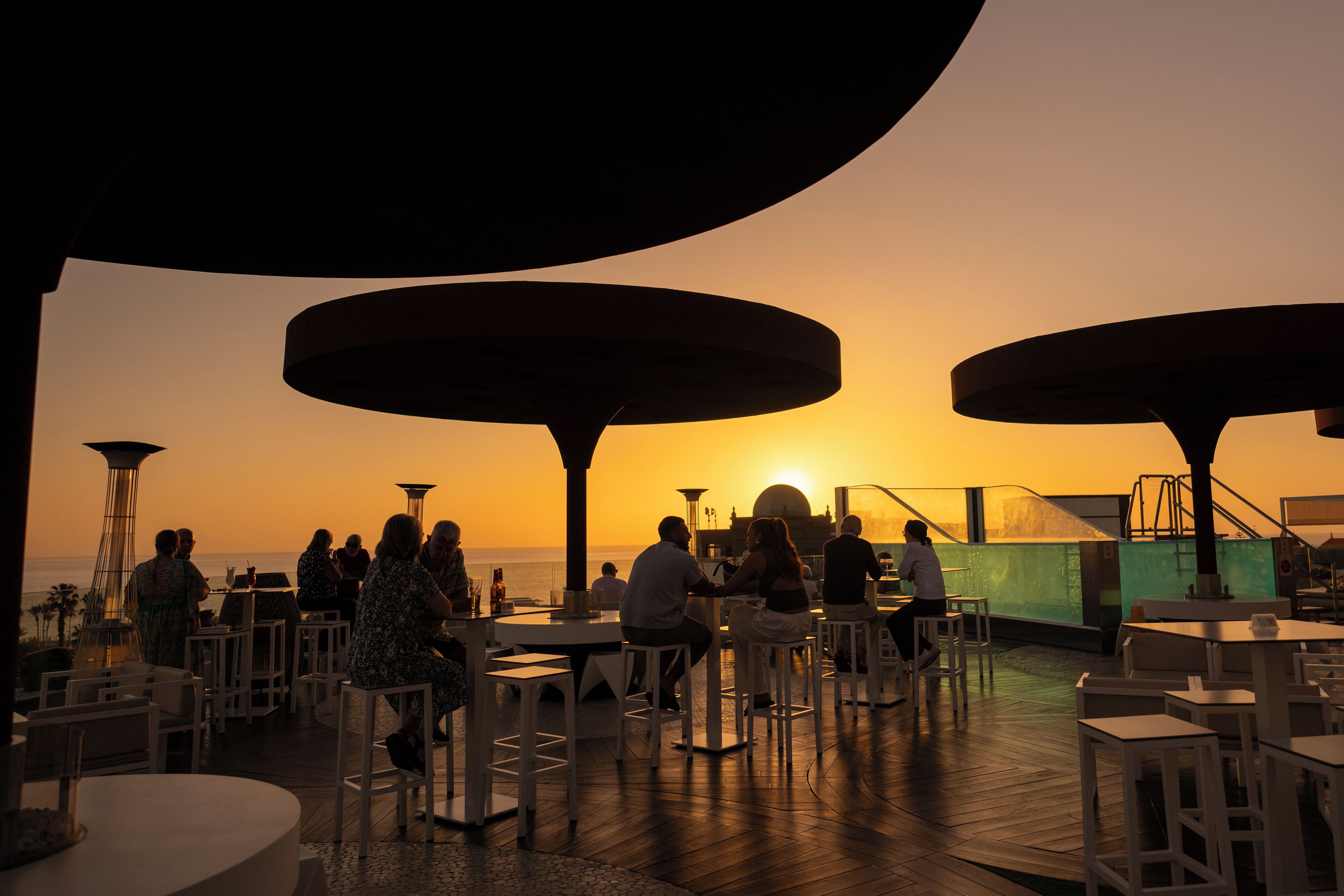 An outdoor dining area with tables and chairs, overlooking the ocean at sunset.