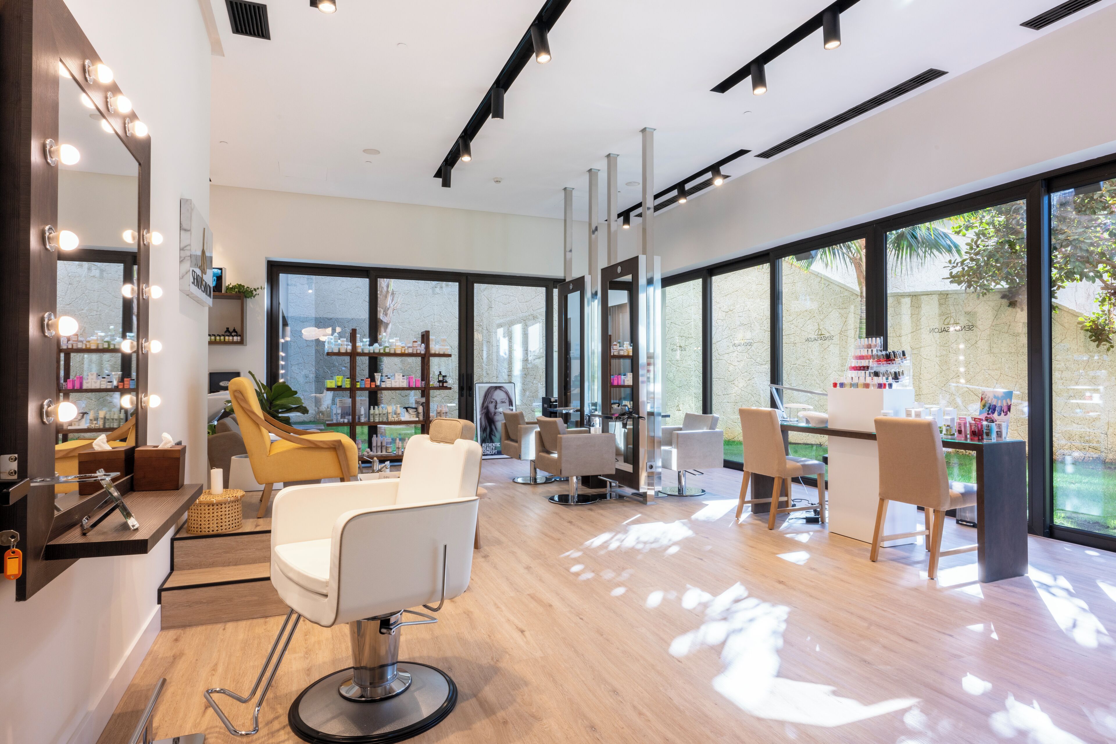 A modern salon with large windows, a mirror with lights, and several chairs.