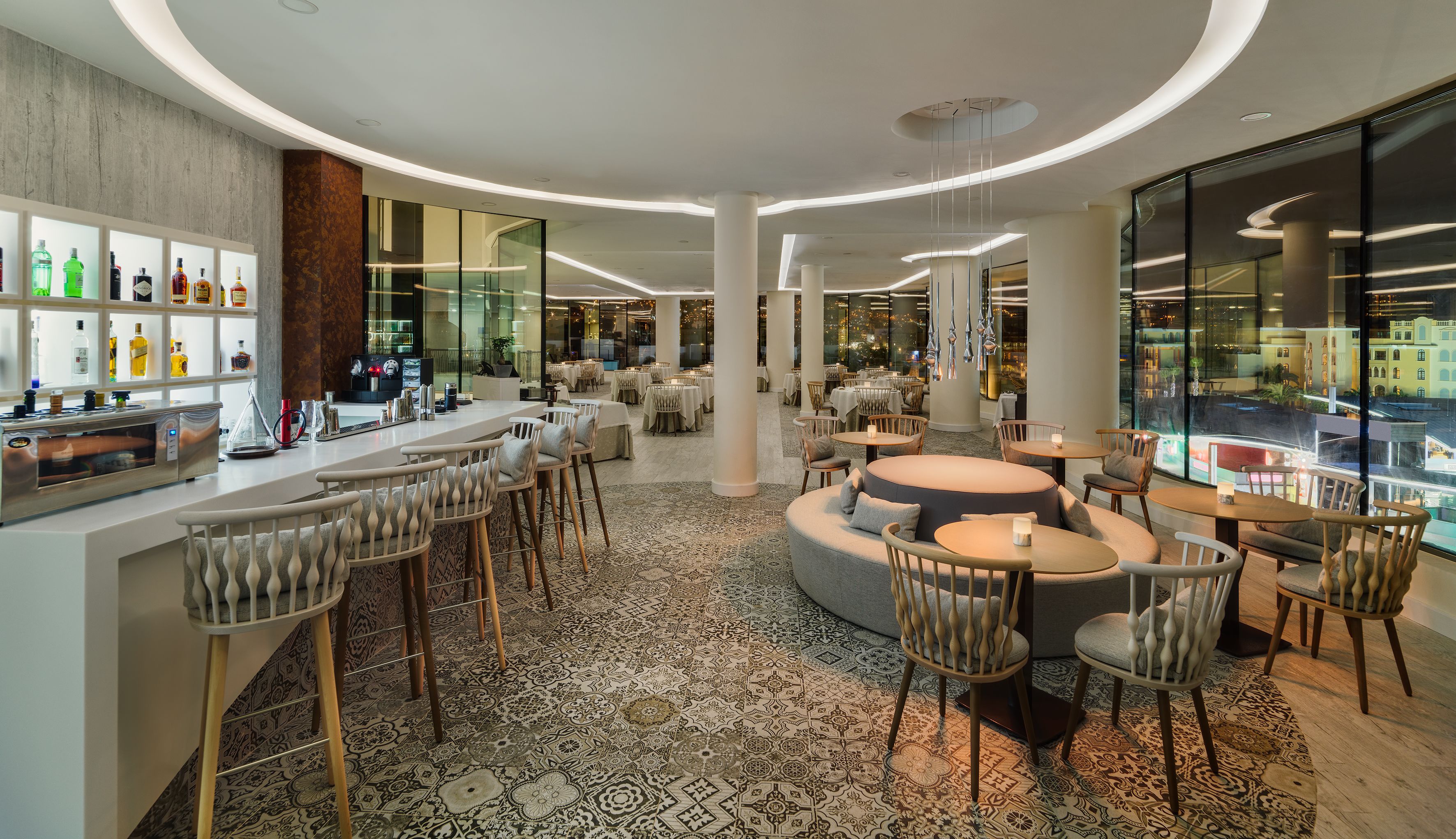 Un espace bar moderne offrant différents types de sièges, notamment des tabourets de bar et des tables rondes, ainsi qu’un présentoir où sont exposées les bouteilles d’alcool.