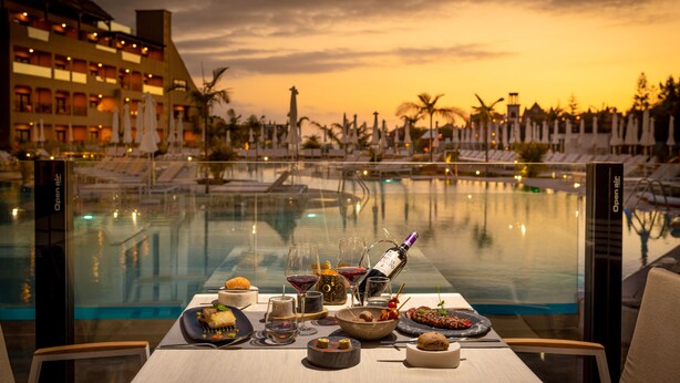 A dining table with a sunset view, a pool, and a hotel building in the background. - GF Victoria (Adeje)