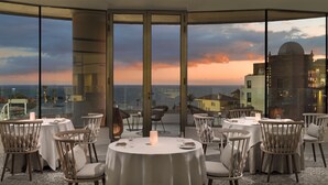 A rooftop dining area with round tables and chairs, large windows offering a city view at sunset. - GF Victoria (Adeje)
