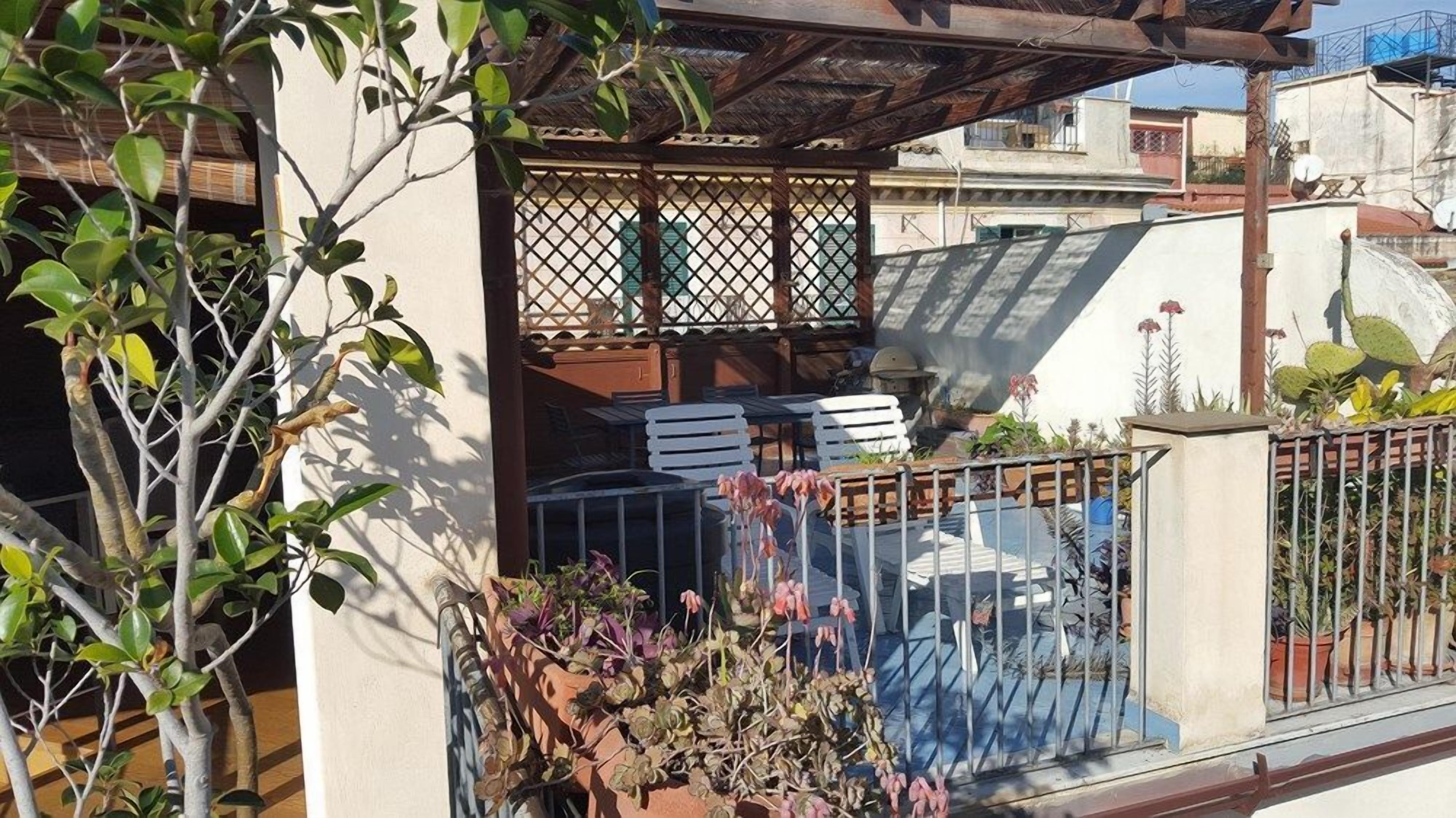 A balcony with a wooden pergola, white chairs, and potted plants.