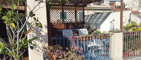 A balcony with a wooden pergola, white chairs, and potted plants.