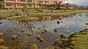 Un edificio moderno con techo plano y ventanas amplias, rodeado por un paisaje natural con un estanque y vegetación escasa.