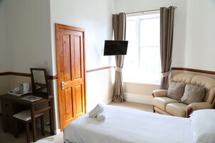 A hotel room with a wooden desk, a chair, a bed, a TV mounted on the wall, and a window with curtains.
