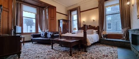A traditional hotel room with a large bed, a sofa, a desk, and a fireplace.