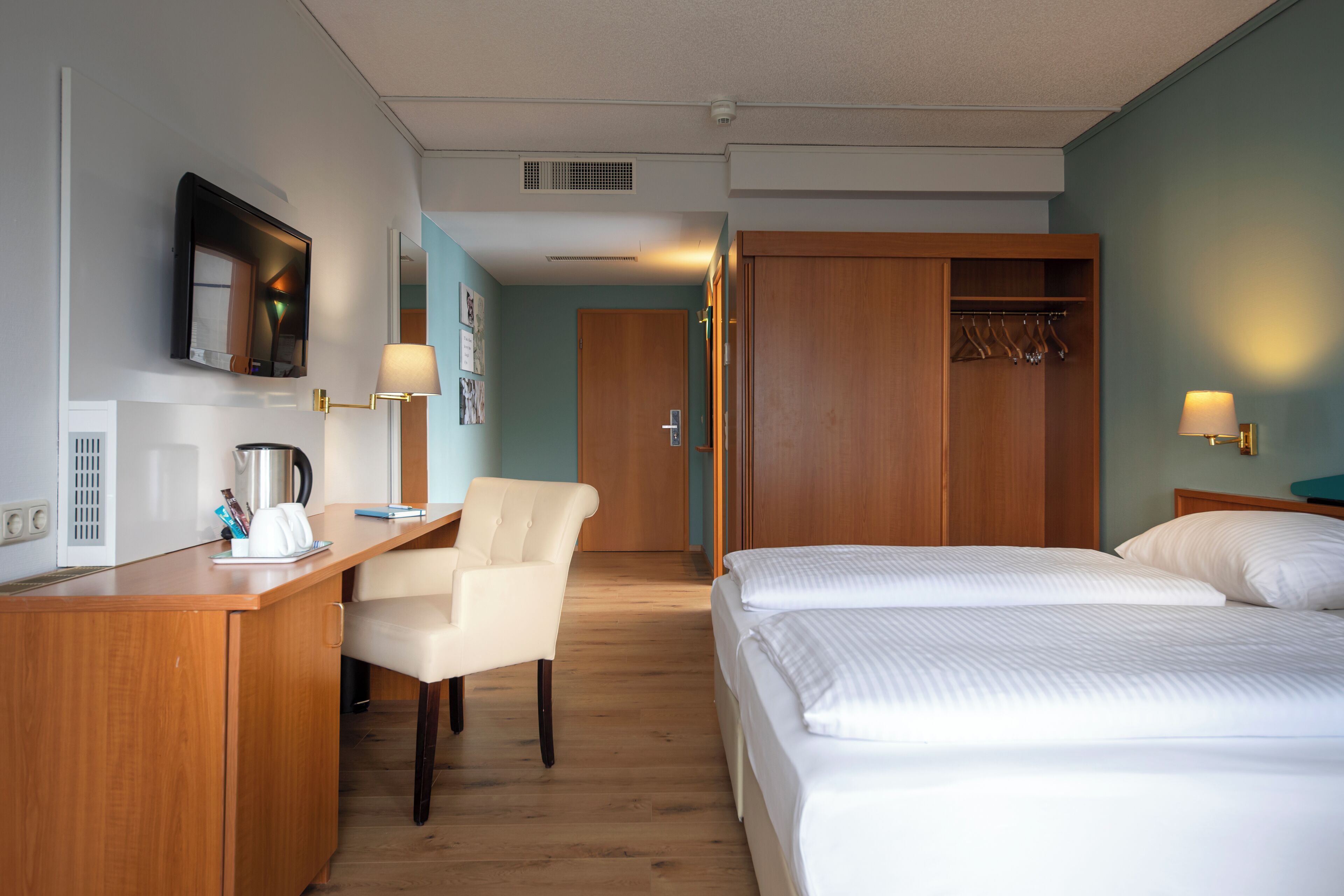 A hotel room with a wooden desk, a chair, a bed with white bedding, a wardrobe, and a television mounted on the wall.