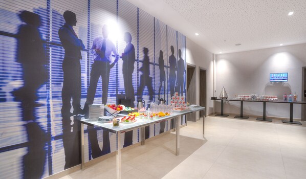 A modern conference room with a long table set for a meeting, a coffee station, and a television screen on the wall.