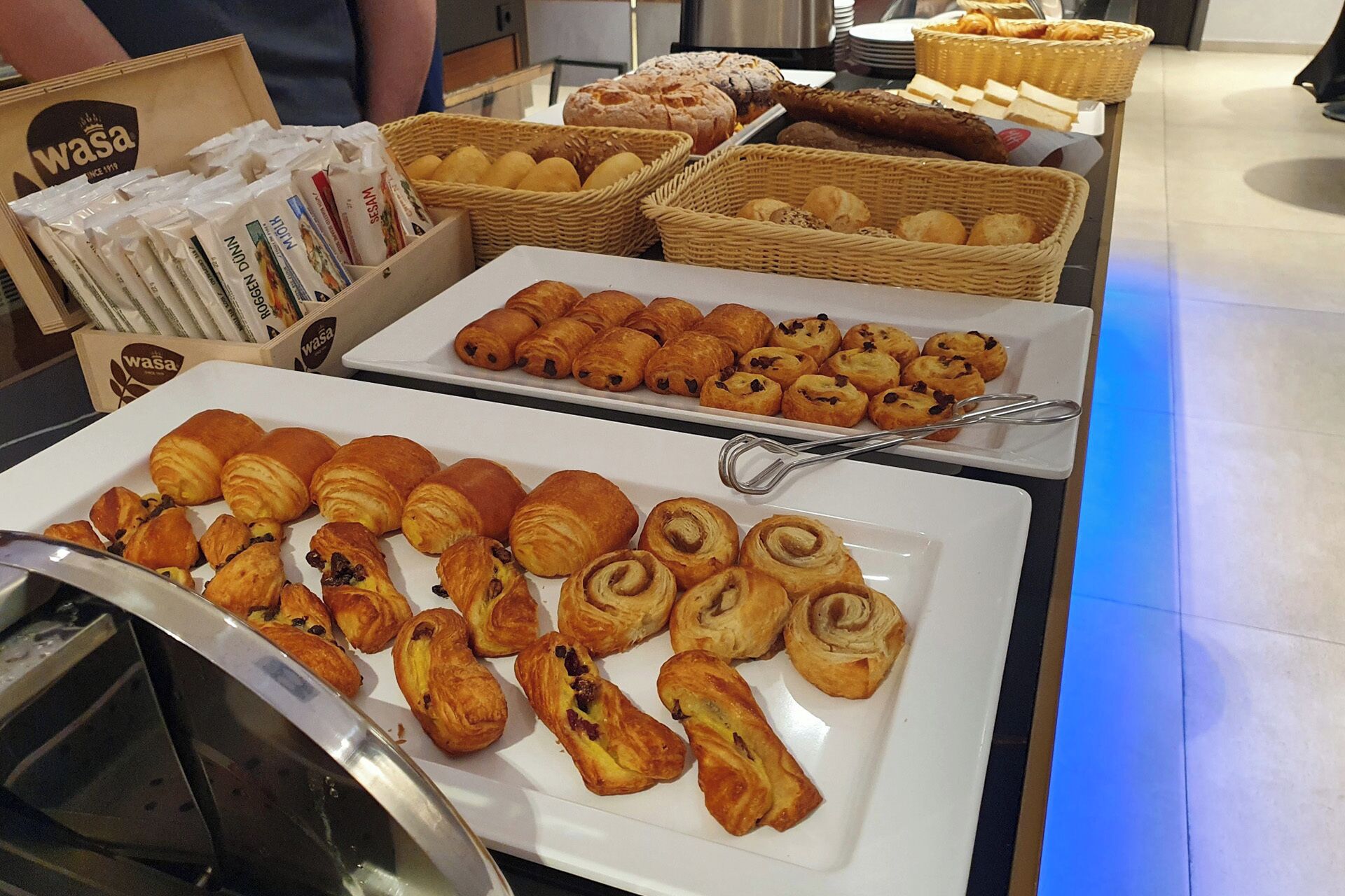 Un bufé con diversas pastas y panes, incluyendo cruasanes y muffins, exhibidos en platos blancos y cestas de mimbre.