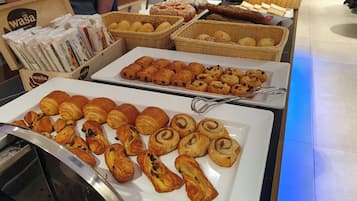 Un bufé con diversas pastas y panes, incluyendo cruasanes y muffins, exhibidos en platos blancos y cestas de mimbre.