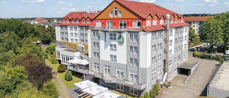 Un hotel de varios pisos con techo rojo, un área de restaurante visible con mesas al aire libre y un estacionamiento.