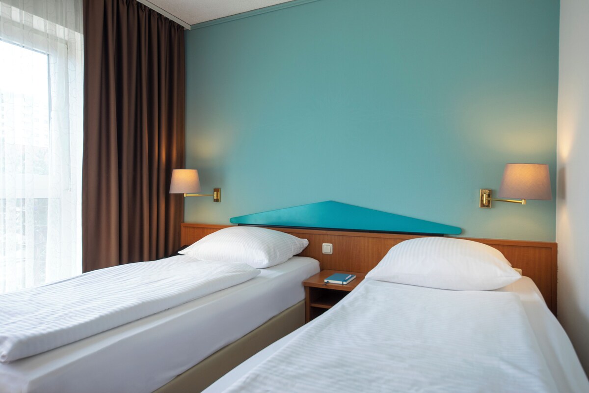 A hotel room with two single beds, a wooden headboard, a nightstand with a lamp, and a view of the city through large windows.
