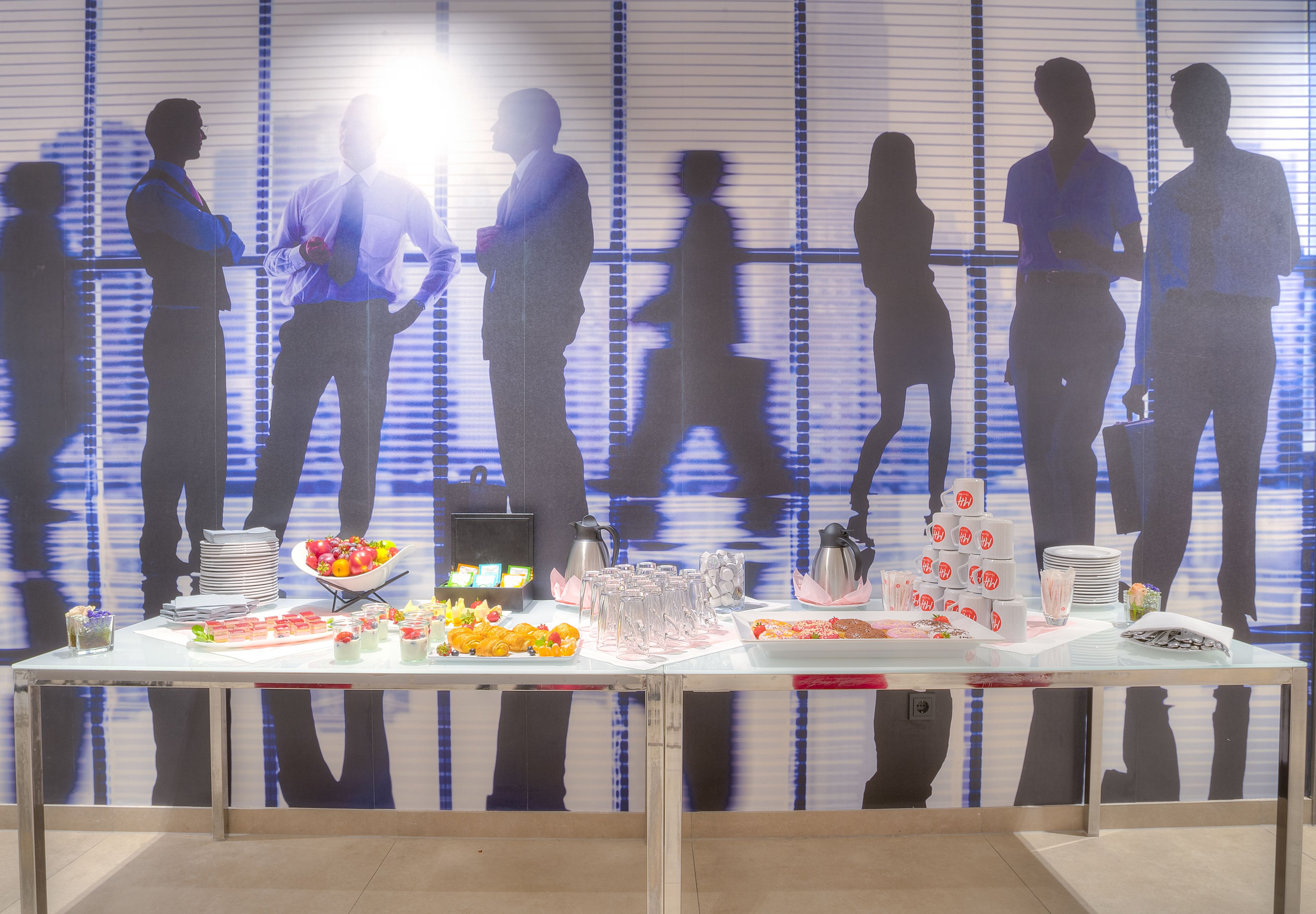 A buffet table with various food items and drinks, set against a backdrop of silhouettes of people.