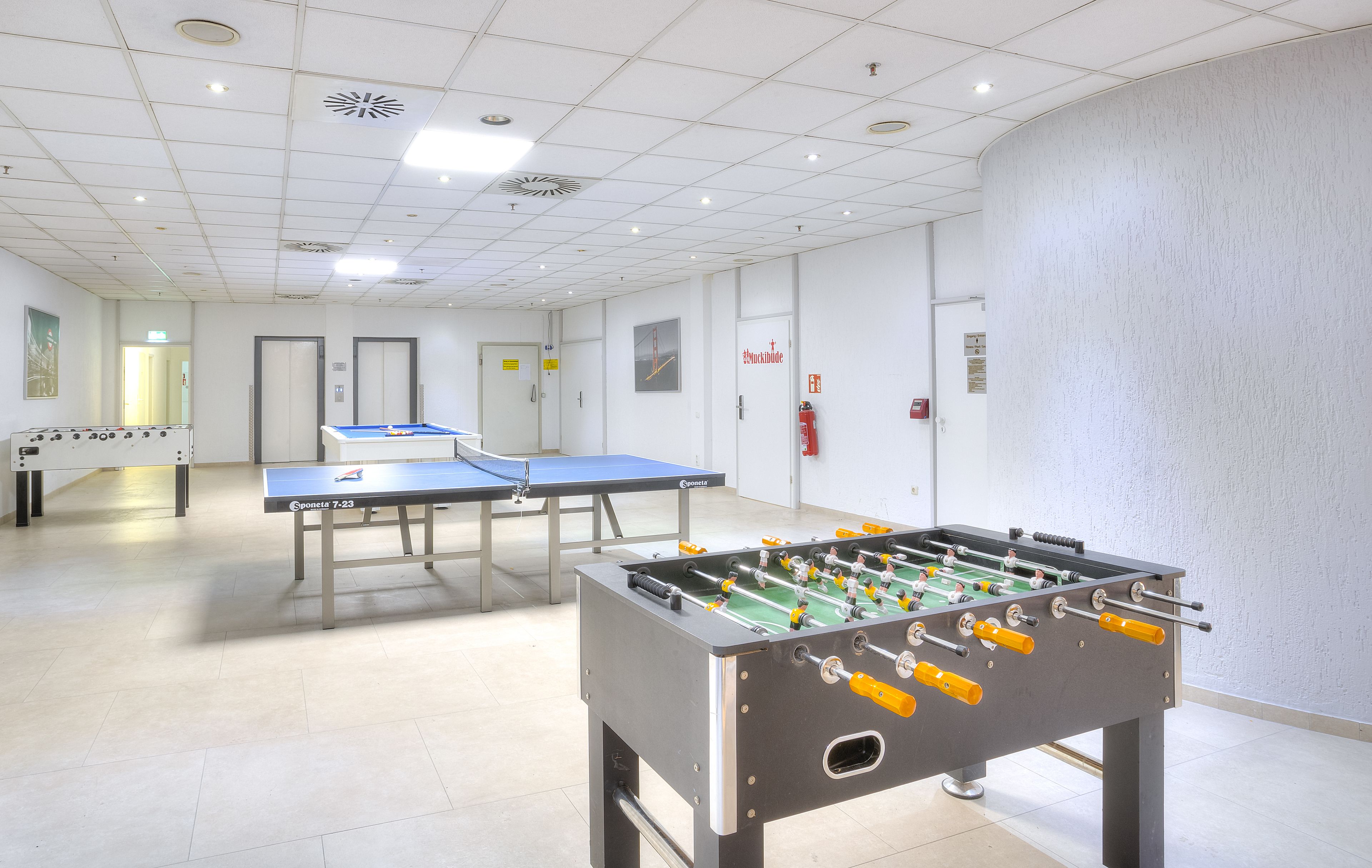 Una habitación con dos mesas de fútbol de mesa y una mesa de ping pong, paredes blancas y piso de baldosas.