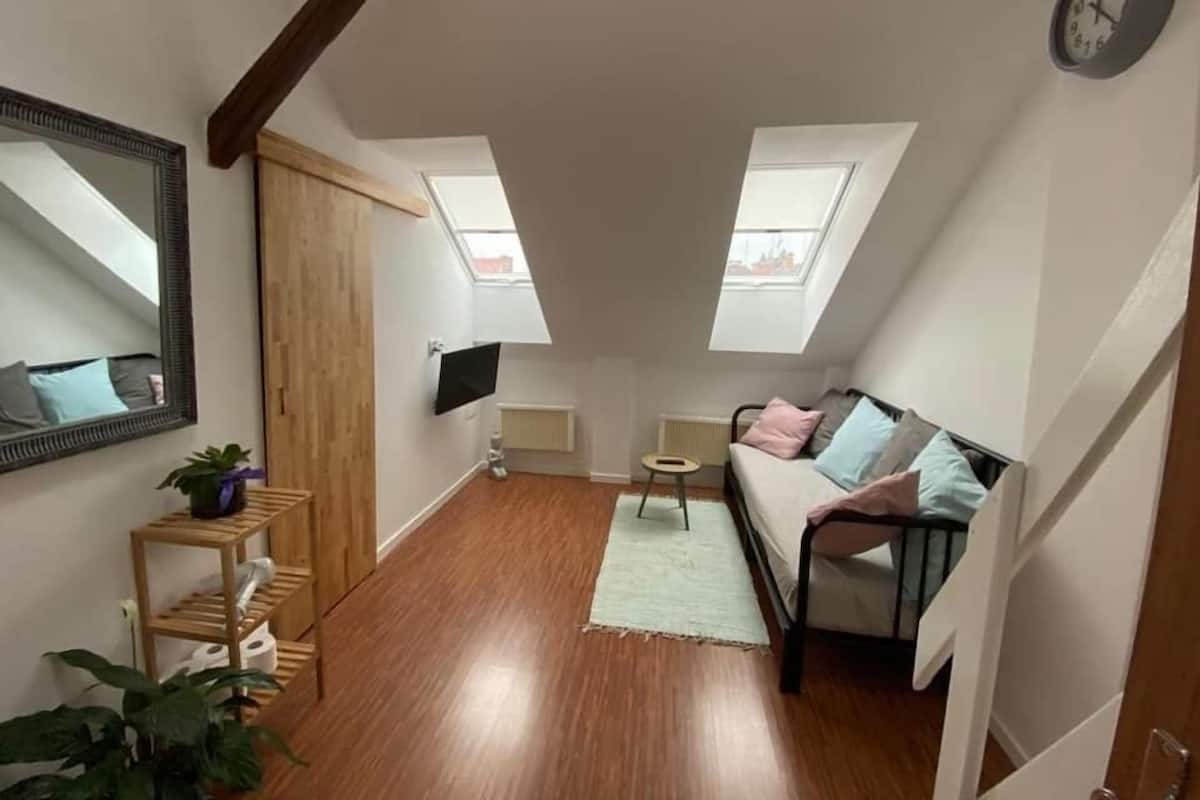 A small, well-lit attic bedroom with a sloped ceiling, a bed, a TV, a small table, and a mirror.