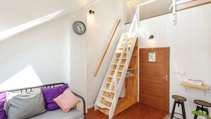 A dorm-style room with a bunk bed, a small desk with a chair, a ladder, a wooden door, and a wall-mounted shelf with a tissue box. - Pension Skylark (Pardubice)