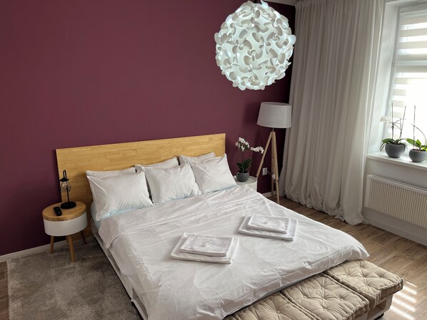 A neatly made bed with white linens, a wooden headboard, and a beige upholstered bench. There is a bedside table with a lamp, a potted plant, and a small decorative item. A unique white pendant light hangs above the bed. - Pension Skylark (Pardubice)