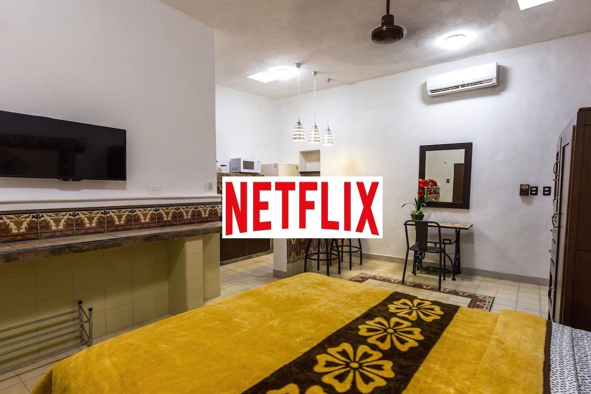 A hotel room with a bed, a TV mounted on the wall, a desk with a chair, and an air conditioning unit.