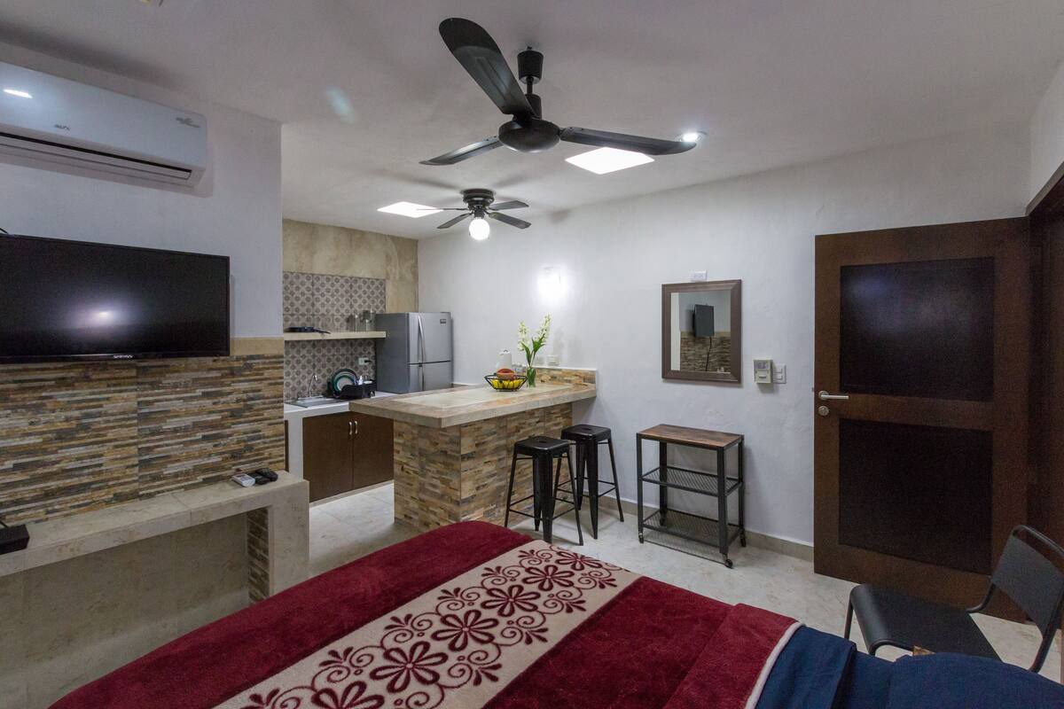 A compact hotel room with a kitchenette, a ceiling fan, a TV, a bed with a red and white patterned bedspread, and a small seating area with a chair and stools.