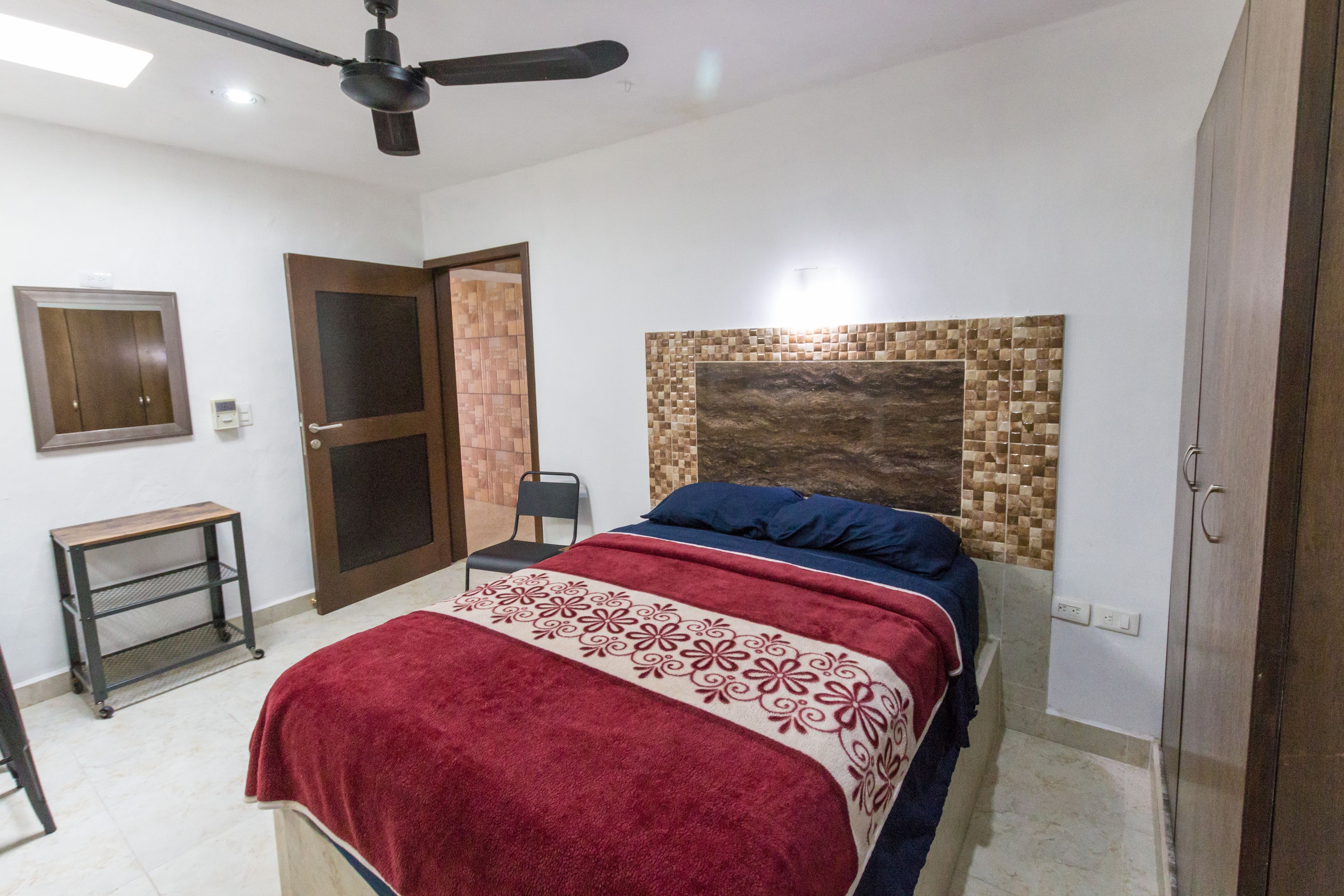 Una camera da letto con un letto, una ventola a soffitto, una porta, un tavolino e una mensola fissata al muro.