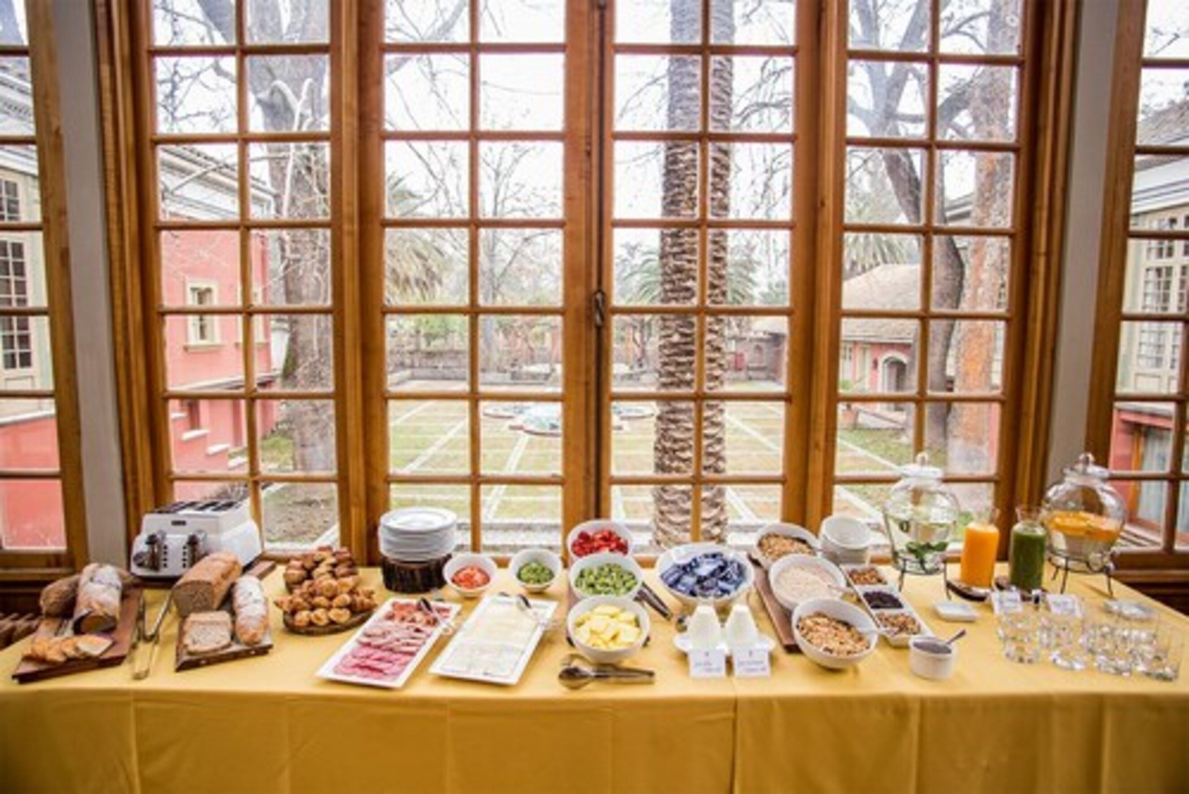 Une table de buffet garnie de divers plats et agrémentée d’un grille-pain, placée devant une fenêtre offrant une vue sur des palmiers et des bâtiments.
