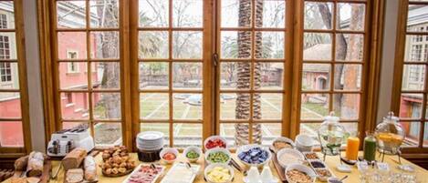 Une table de buffet garnie de divers plats et agrémentée d’un grille-pain, placée devant une fenêtre offrant une vue sur des palmiers et des bâtiments.
