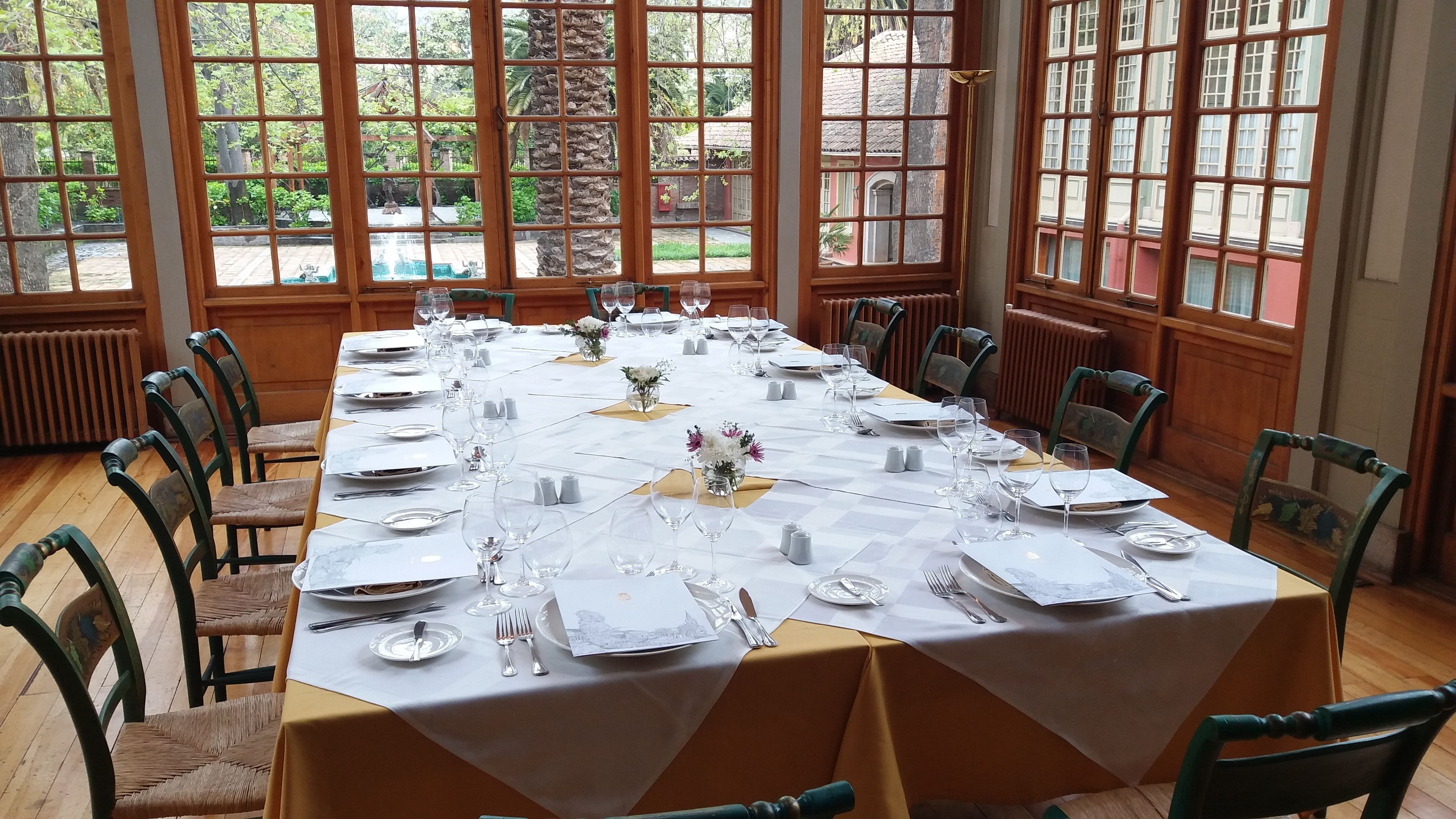 Una larga mesa de comedor dispuesta para una comida formal, con sillas alrededor, grandes ventanales con vistas a zonas verdes y edificios, y un piso de madera.