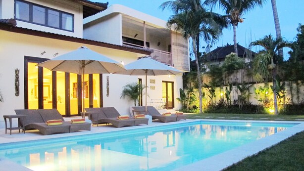 A swimming pool with lounge chairs and umbrellas, a two-story building with balconies, and palm trees in the background. - Aisha Family Villas-5Bedroom Private Pool (Seminyak)