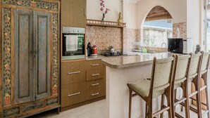 A kitchen with a large wooden cabinet, a built-in oven, a brick backsplash, and a bar area with stools. - Aisha Family Villas-5Bedroom Private Pool (Seminyak)