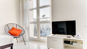 Una sala de estar moderna con un ventanal grande, un televisor de pantalla plana sobre un mueble blanco, una silla de mimbre con cojín naranja y un sofá blanco.