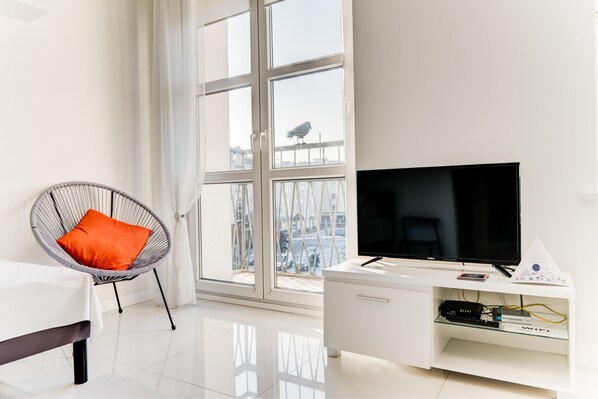A modern living room with a large window, a flat-screen TV on a white cabinet, a wicker chair with an orange cushion, and a white sofa.