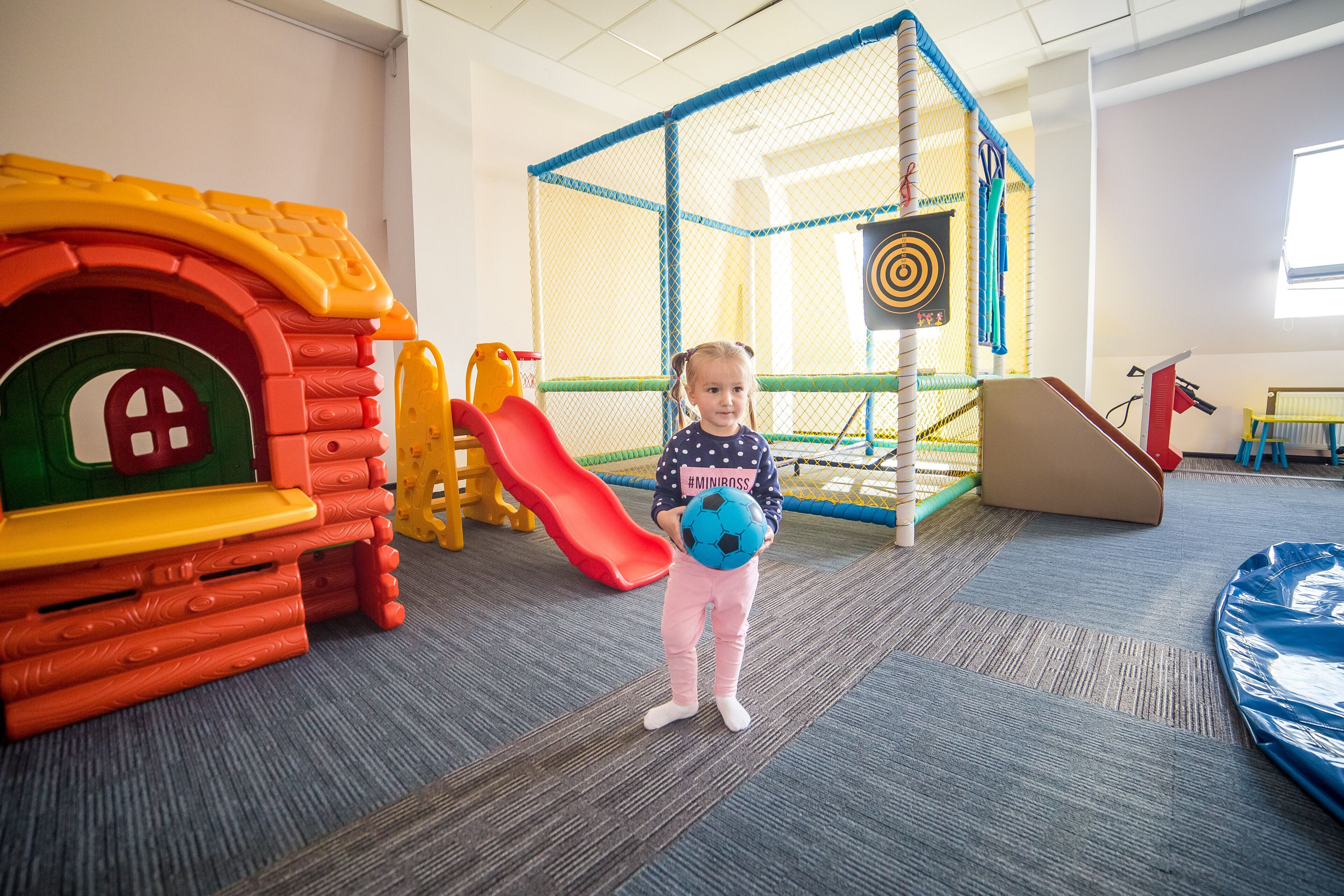 A child in a play area with a slide, climbing wall, and netted play structure.