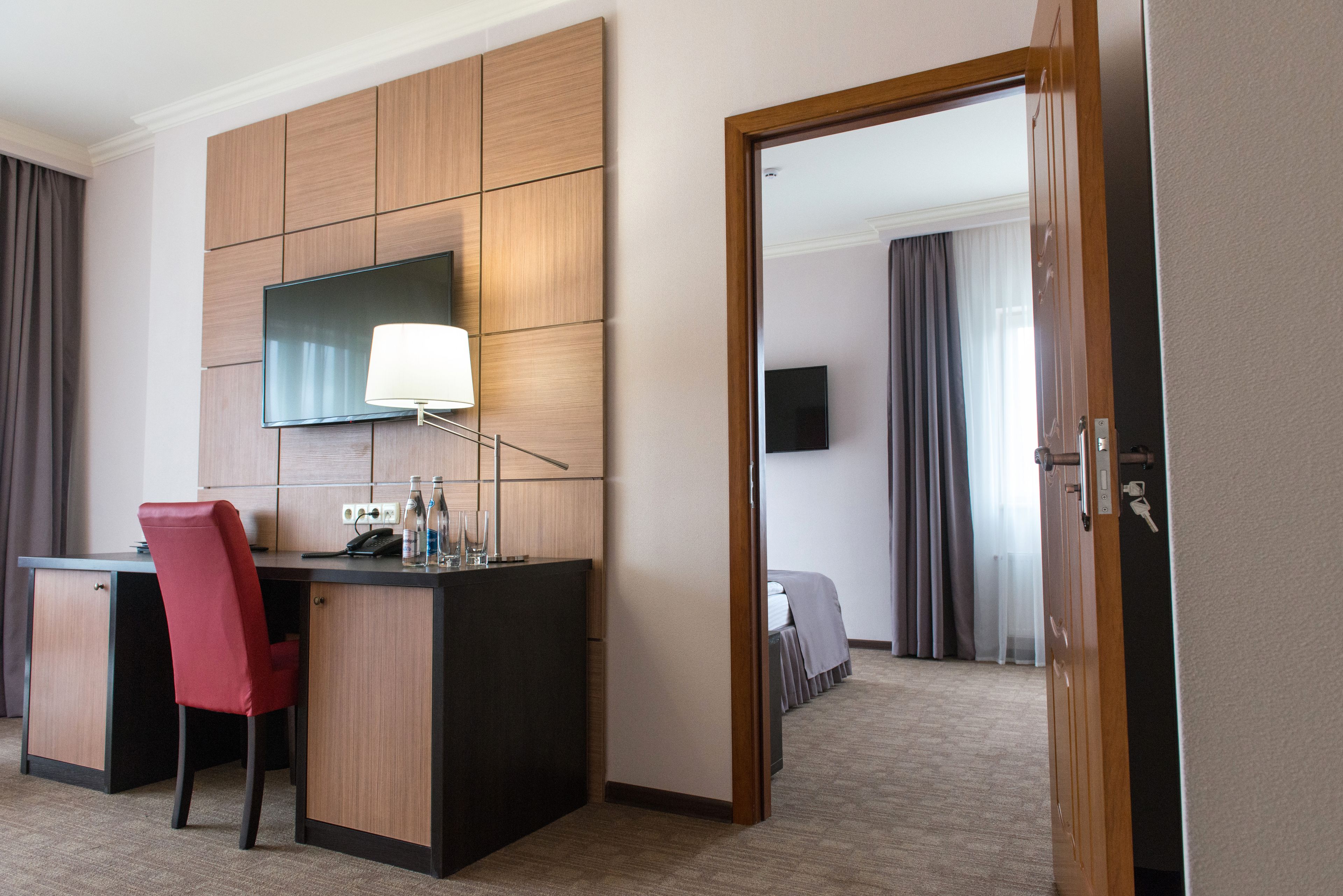 Habitación de hotel con escritorio, una silla roja, televisor de pantalla plana montado en la pared, mesita de noche con lámpara y cama con colcha blanca.