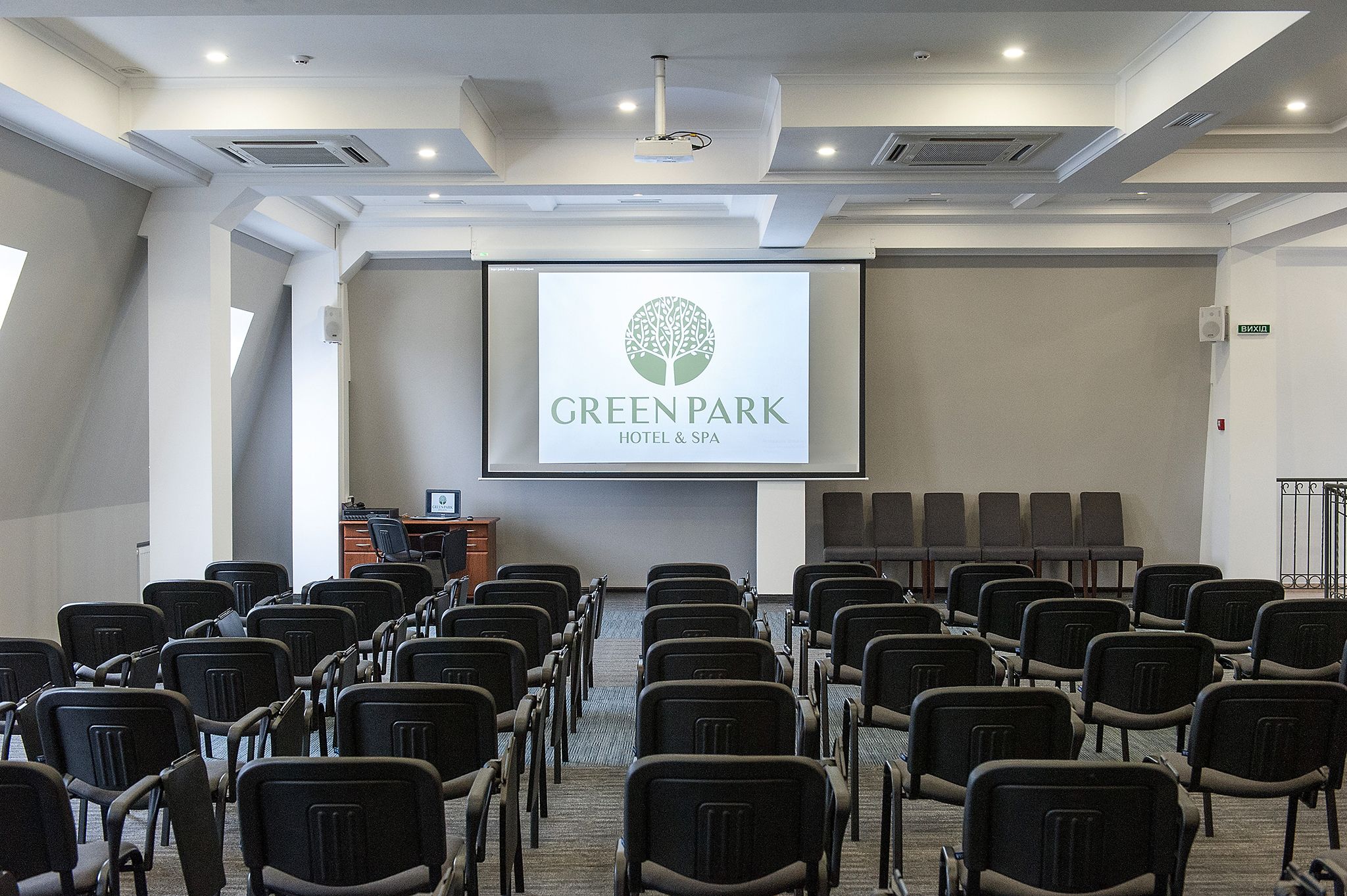 Une salle de conférence avec des rangées de chaises, un écran de projection affichant le logo de l’hôtel et du spa GreenPark, et un pupitre à l’avant.