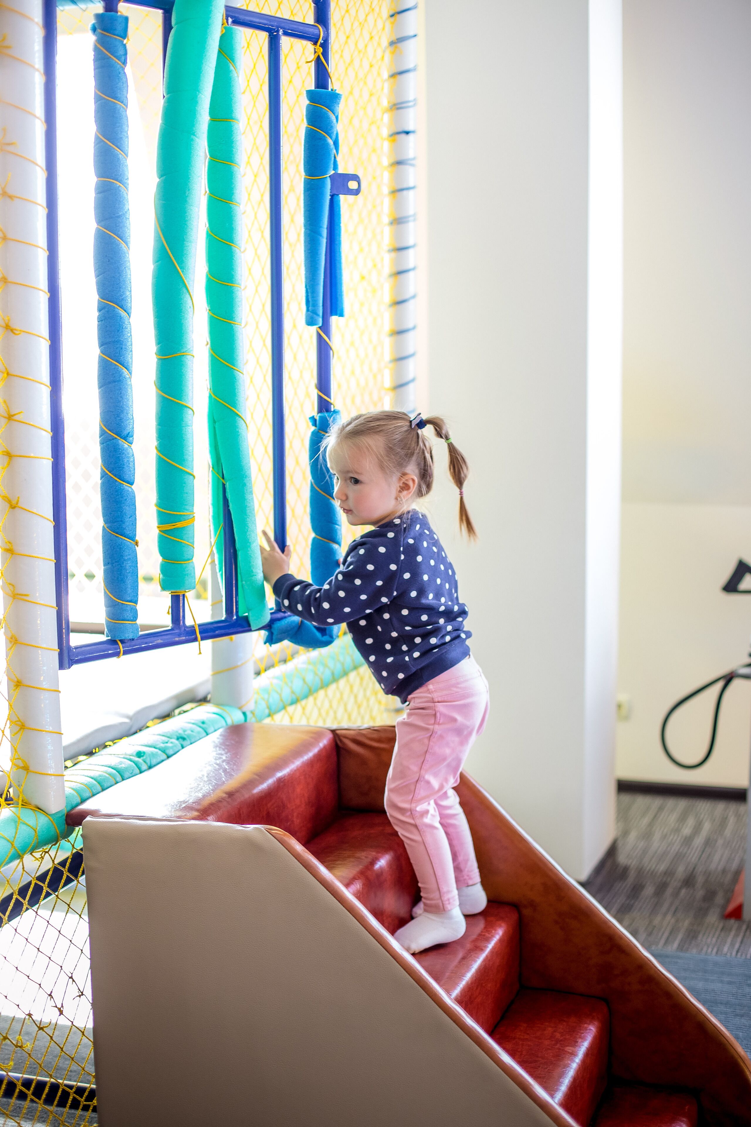 A child climbing a red staircase in a play area with netted walls and padded steps.