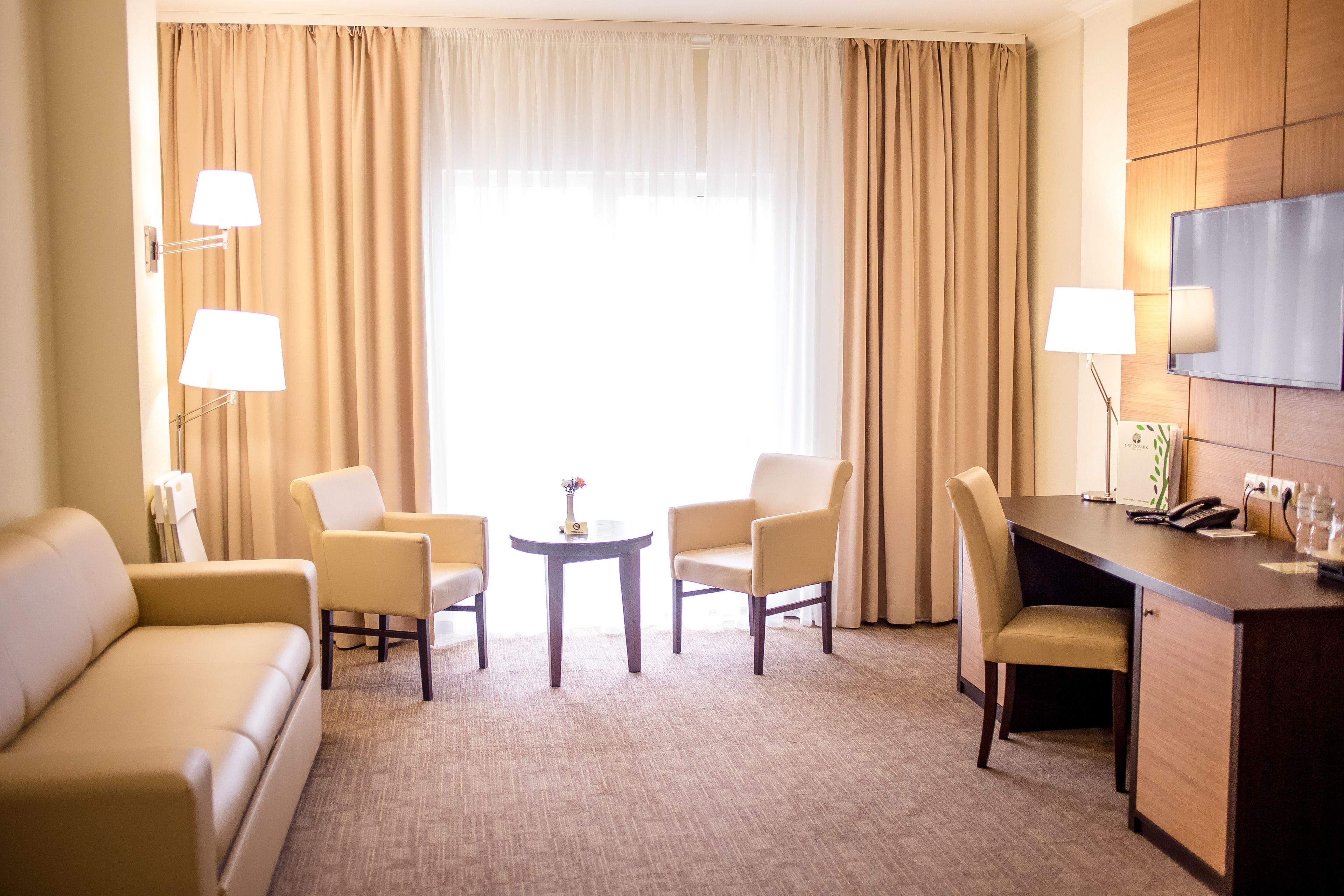 A hotel room with a sofa, two armchairs, a small round table, a desk with a lamp, and a TV on the wall.